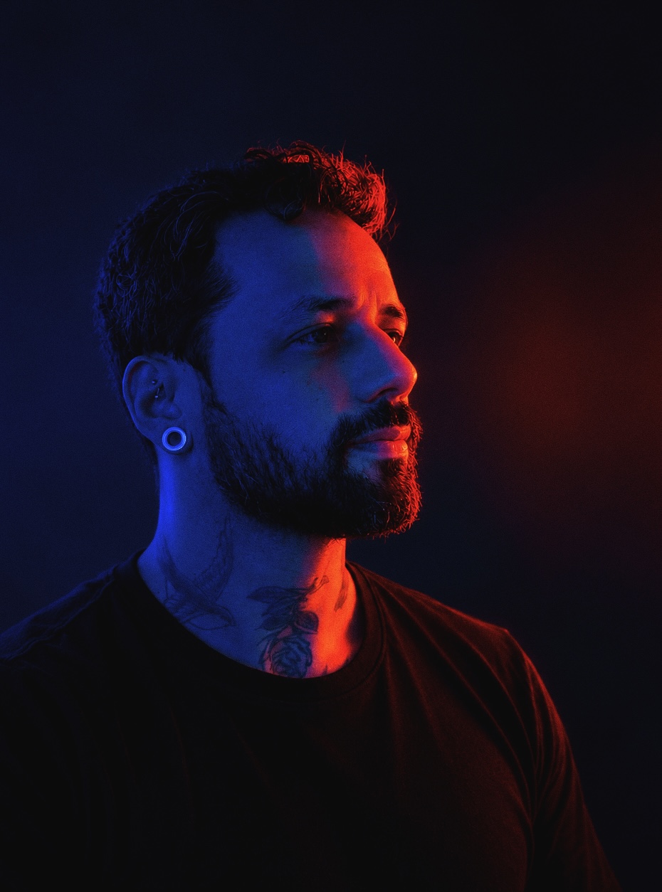 Patric Teixeira in profile, lit blue on one side and red on the other, with curly hair, a beard, an ear gauge, neck tattoos and a black T-shirt.